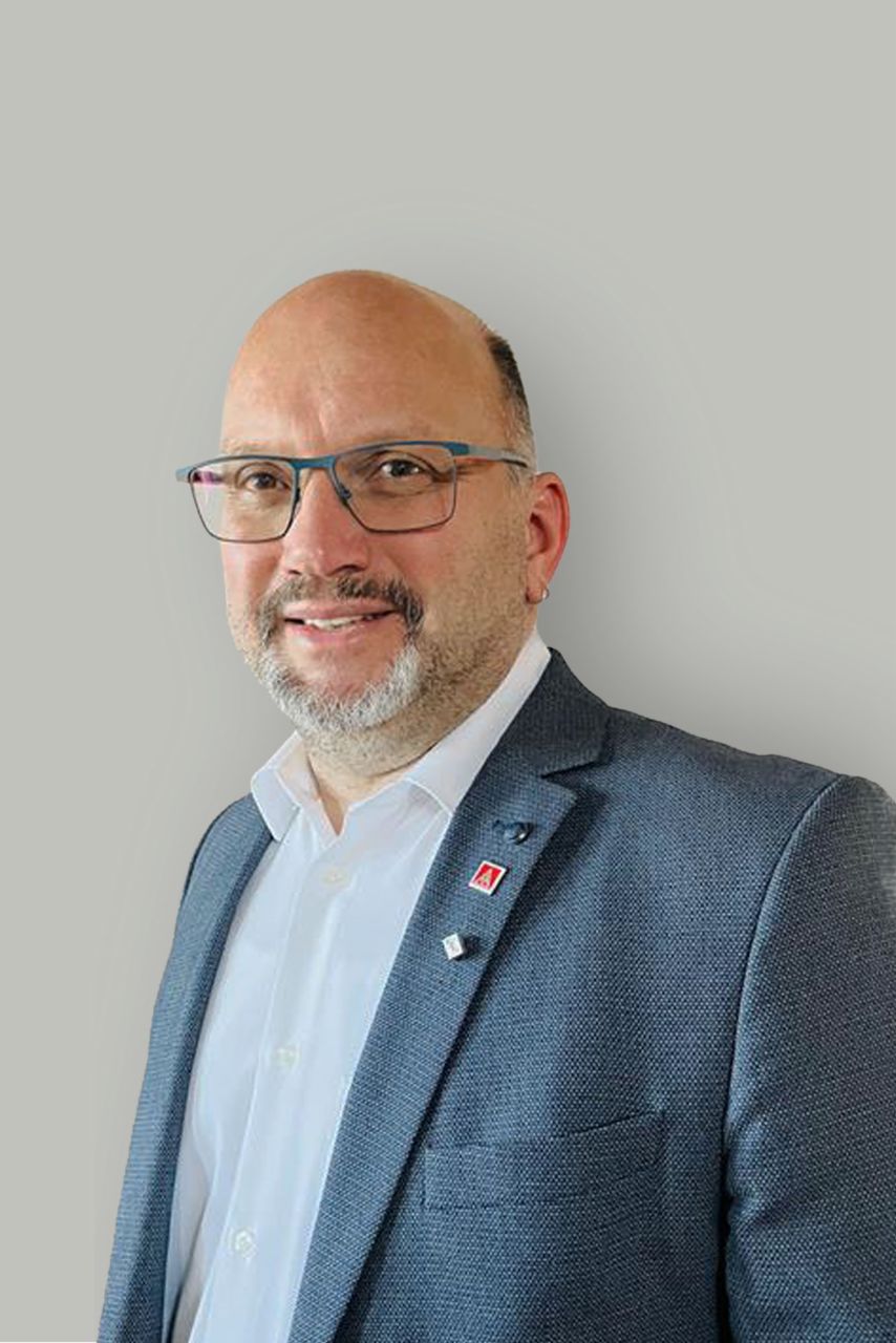 Ein Mann in einem Anzug steht vor einen grauen Wand und lächelt in die Kamera