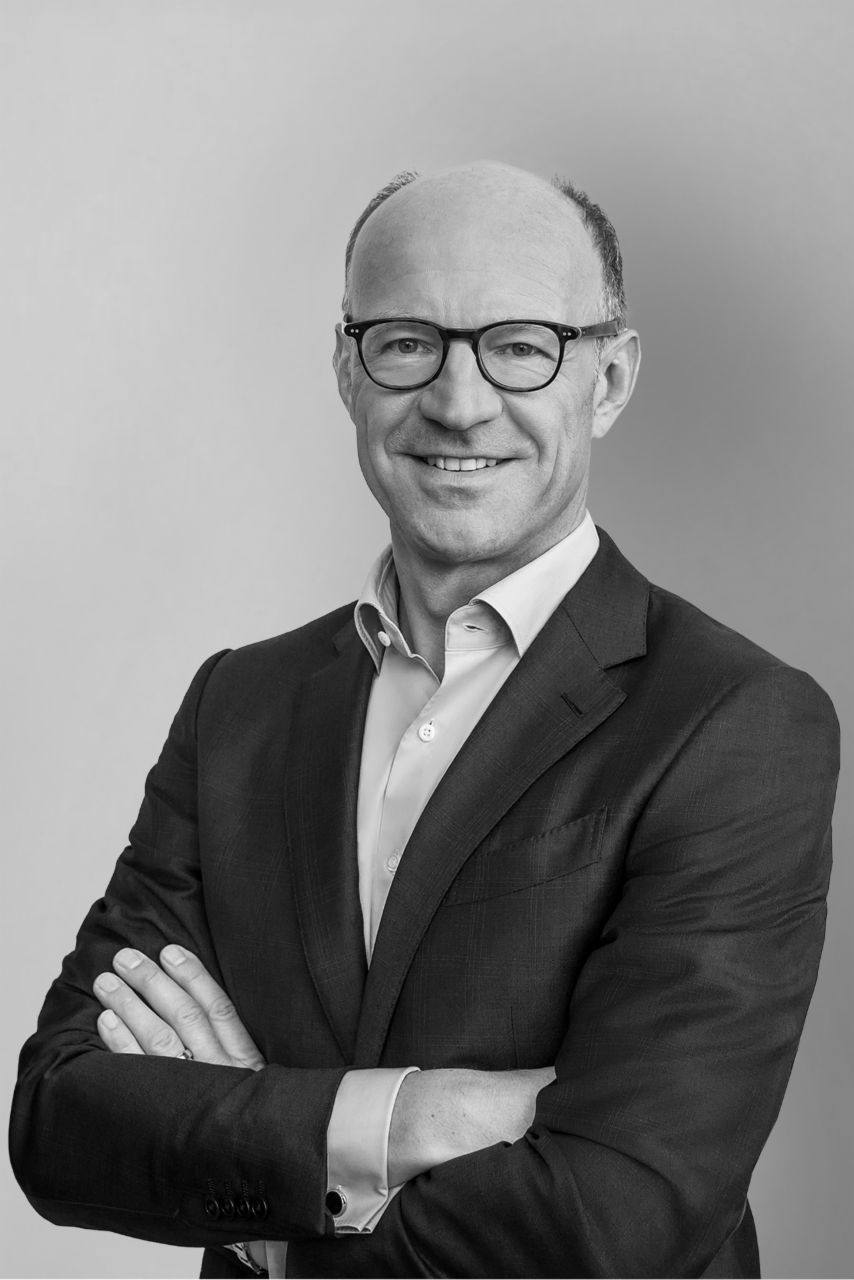 Ein Mann mittleren Alters im blauen Anzug und hellblauen Hemd steht mit verschränkten Armen vor einer Betonwand. Er trägt eine Brille und lächelt in die Kamera.