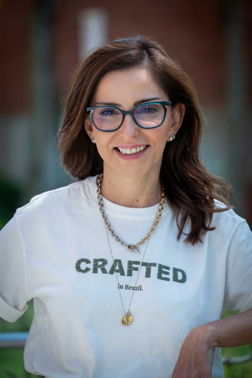 Patricia Acioli standing outdoors in front of a softly blurred background featuring buildings and greenery. Patricia Acioli is wearing glasses, a light-colored T-shirt displaying the text “CRAFTED in Brazil.”, and two necklaces. The image is a close portrait showing the upper body against a shallow-depth-of-field background.