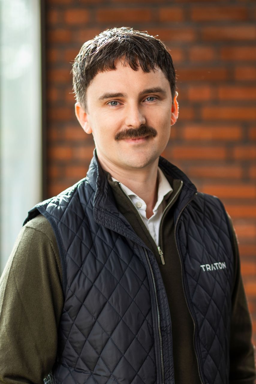 Joakim Dahlborn, Product Strategy Manager at TRATON Group, standing in front of a brick wall with a green sweater and black vest with the TRATON logo on the right side. 