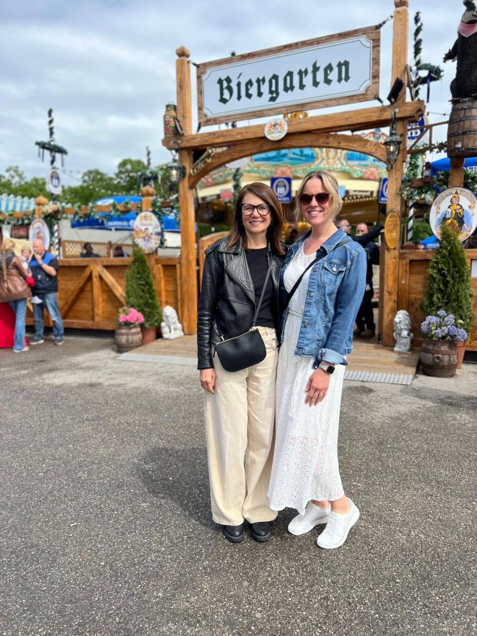 Jana und ihre Freundin stehen vor einem Biergarten-Schild und lächeln in die Kamera. Im Hintergrund sind Menschen und die Dekoration eines Biergartens zu sehen.