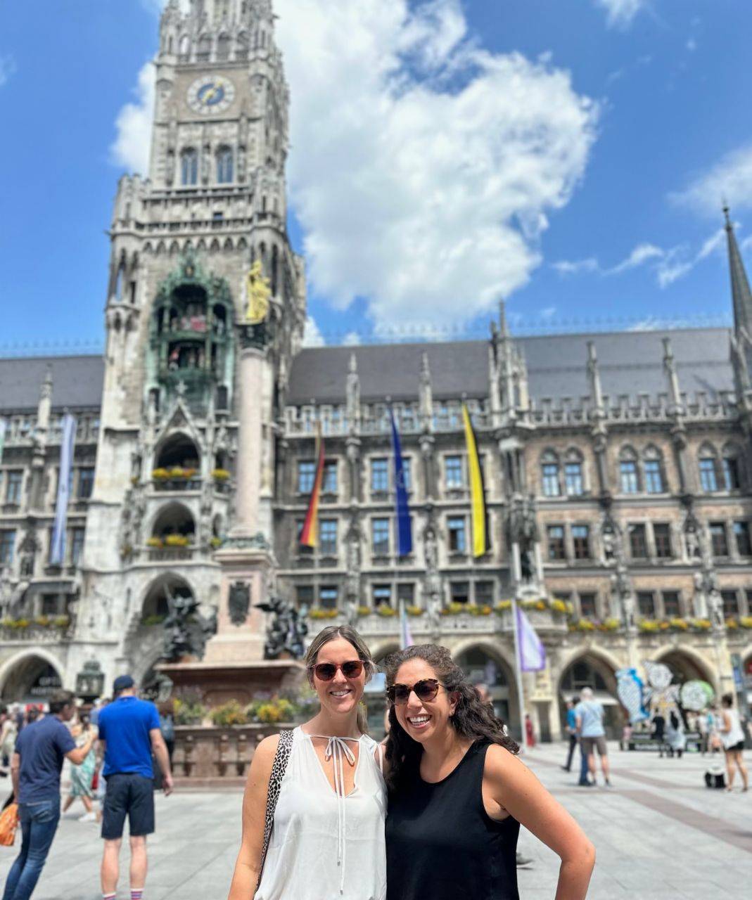 Jana and a friend stand in front of the New Town Hall in Munich, smiling at the camera. It is a sunny day, and other tourists and the magnificent building of the town hall are visible in the background. 