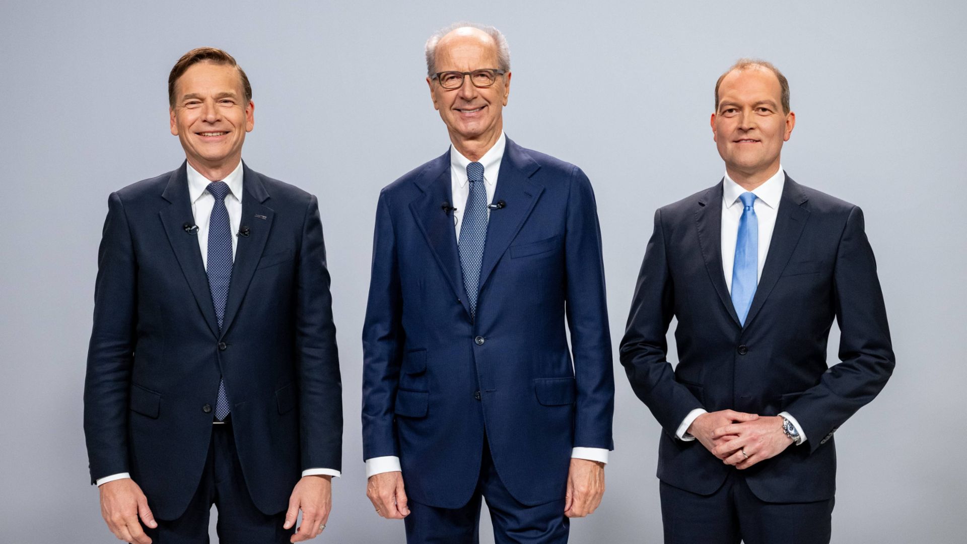  Drei Männer im Anzug stehen nebeneinander vor einer grauen Wand und lächeln. 