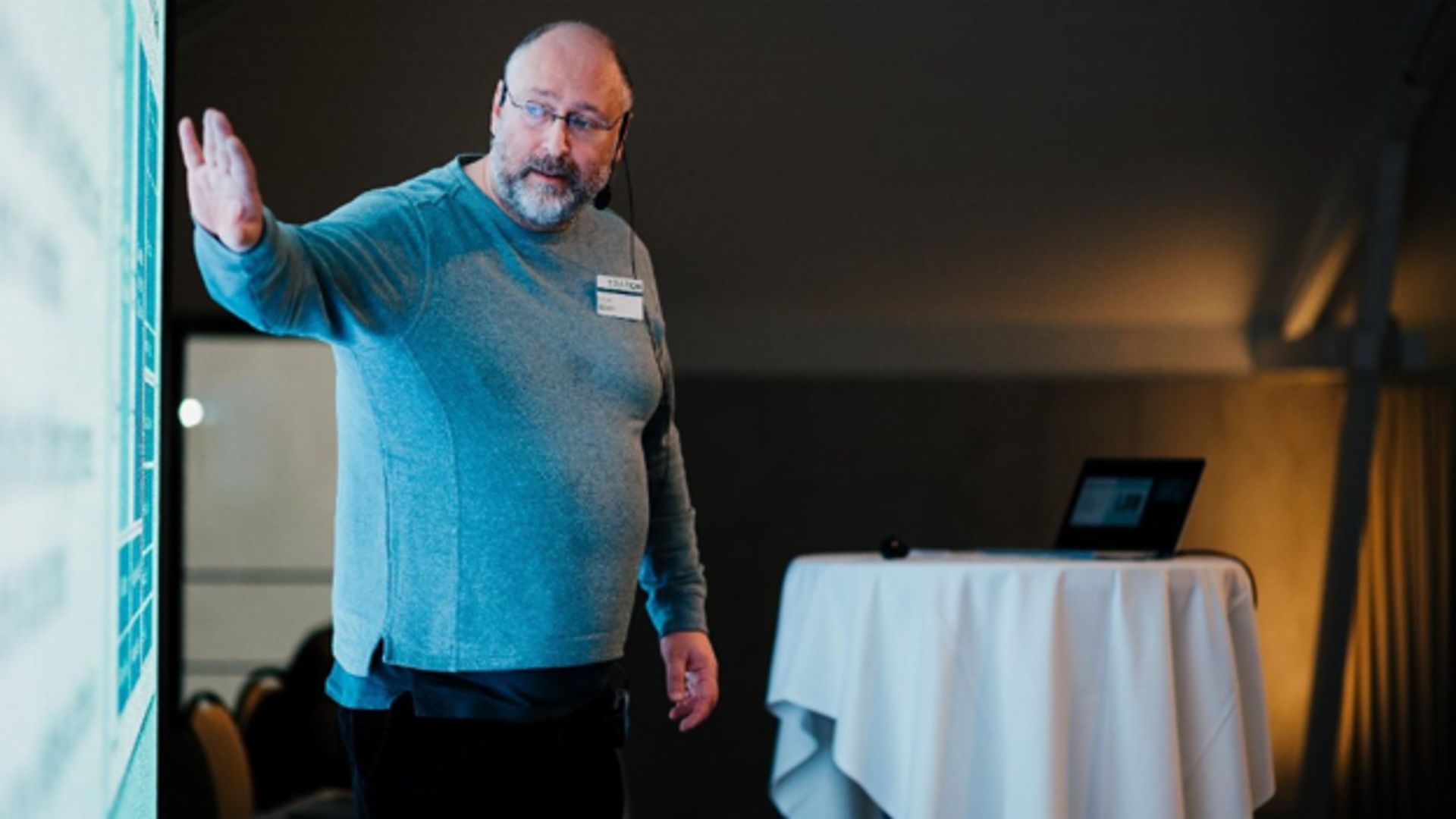 A man in a gray sweater giving a presentation in front of a screen, with a laptop and round table in the background.