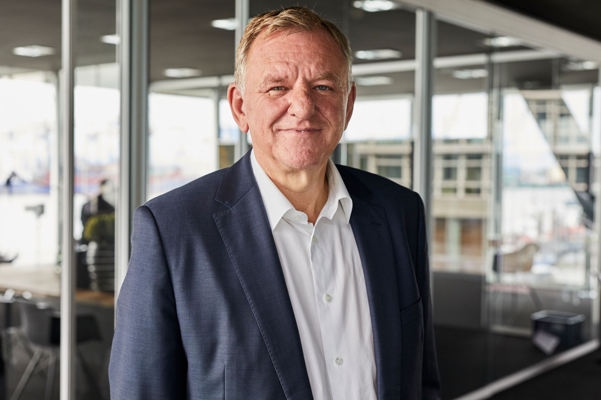 Andreas Renschler im Profil, steht in einem weißen Hemd und blauem Sakko vor einer Wand aus großen Fenstern
                 