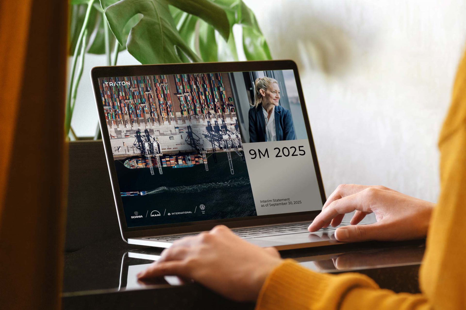A person in a yellow sweater is using a laptop on a dark table. The laptop screen displays a business presentation titled “9M 2025 – Interim Statement as of September 30, 2025” from TRATON Group. The screen shows a container ship seen from above, a portrait of a smiling businesswoman, and logos of TRATON’s brands, including Scania, MAN, International, and Volkswagen Truck & Bus.