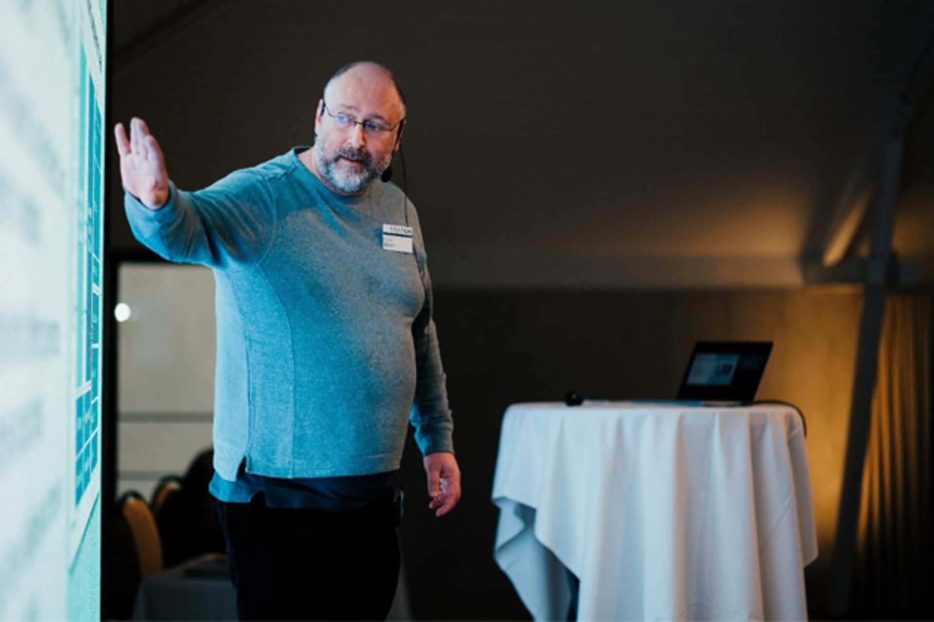 A man in a gray sweater giving a presentation in front of a screen, with a laptop and round table in the background.