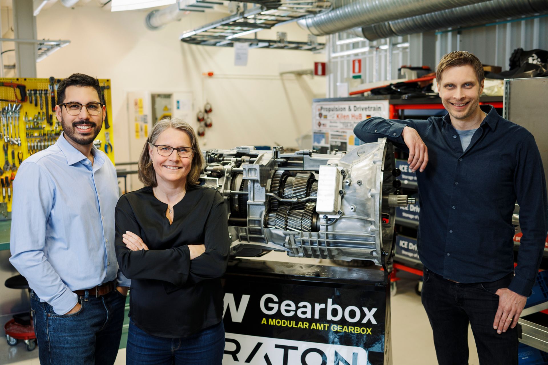 Drei Personen stehen in einer technischen Werkstatt neben einem großen Getriebe mit der Aufschrift „TRATON Gearbox – A modular AMT gearbox“. Von links nach rechts: Markus Granström (mit Brille), Karin Elfgren (mittig) und Peter Daelander (rechts), die Teil des Teams waren, das das gemeinsame Getriebe bei International umgesetzt hat. Im Hintergrund sind Werkzeuge, technische Geräte und Elemente einer Industrieumgebung zu sehen.
