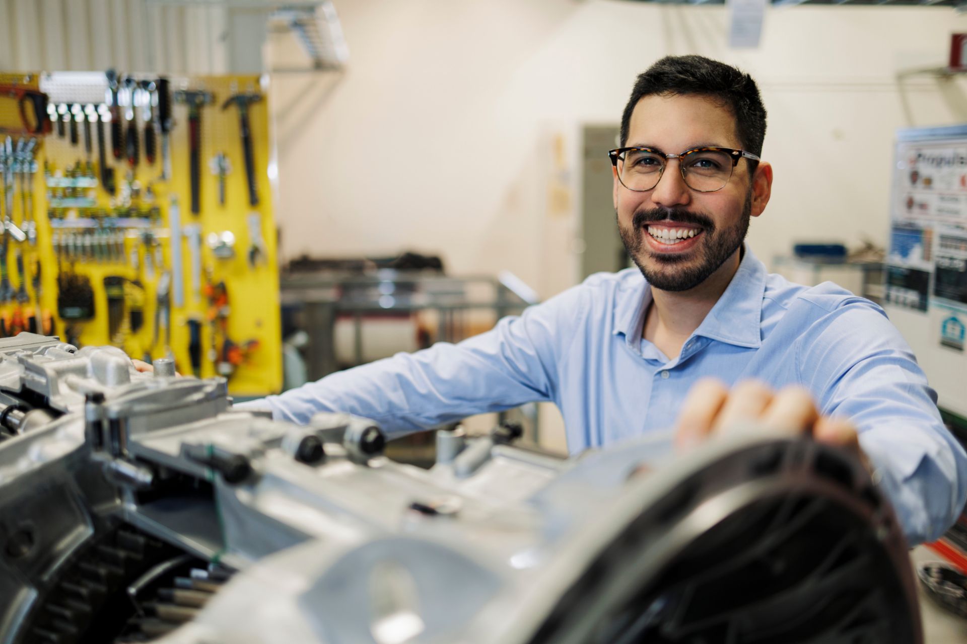 Markus Granström steht in einer Werkstattumgebung und arbeitet an einem großen mechanischen Getriebebauteil, während Markus Granström darauf zeigt. Im Hintergrund sind Werkzeuge an einer gelben Wandtafel sowie weitere industrielle Geräte und Elemente einer Werkstatt zu sehen.

