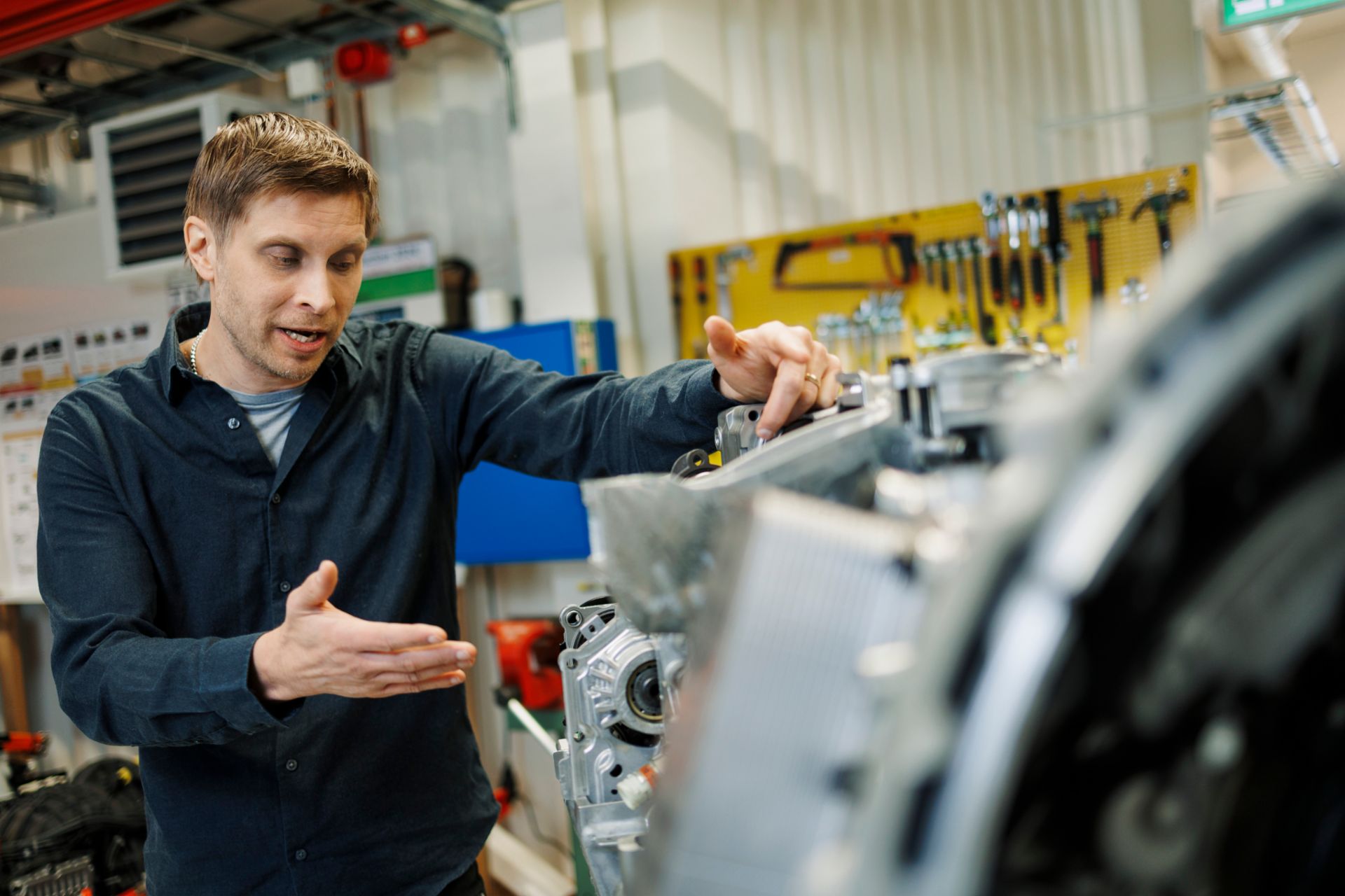 Peter Daelander steht in einer Werkstattumgebung und arbeitet an einem großen mechanischen Getriebebauteil, während er darauf zeigt. Im Hintergrund sind Werkzeuge an einer gelben Wandtafel sowie weitere industrielle Geräte und Elemente einer Werkstatt zu sehen.
