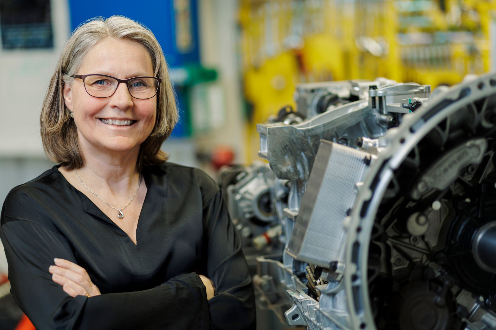 Karin Elfgren steht in einer Werkstattumgebung mit technischen Geräten und Werkzeugen im Hintergrund, zur Kamera ausgerichtet. Die Umgebung umfasst industrielle Elemente wie Maschinen, Werkbänke und geordnetes Werkzeug.