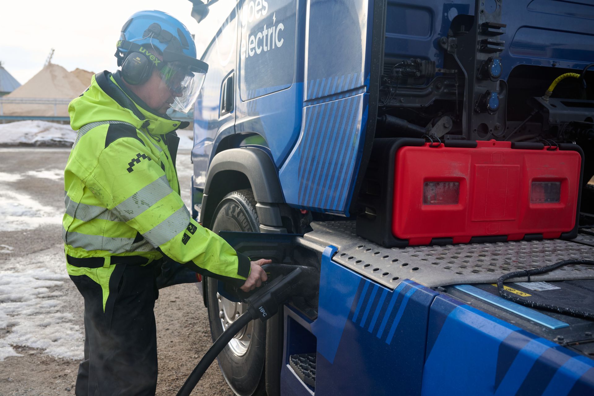 Nahaufnahme eines Arbeiters in vollständiger Sicherheitsausrüstung (Warnjacke, Helm mit Visier und Gehörschutz), der einen HPC-Ladestecker in die Ladebuchse eines blauen Lastwagens steckt. Auf dem LKW ist der Schriftzug „goes electric“ zu sehen. Im Hintergrund ist ein industrielles Gelände mit Schneeresten erkennbar.
