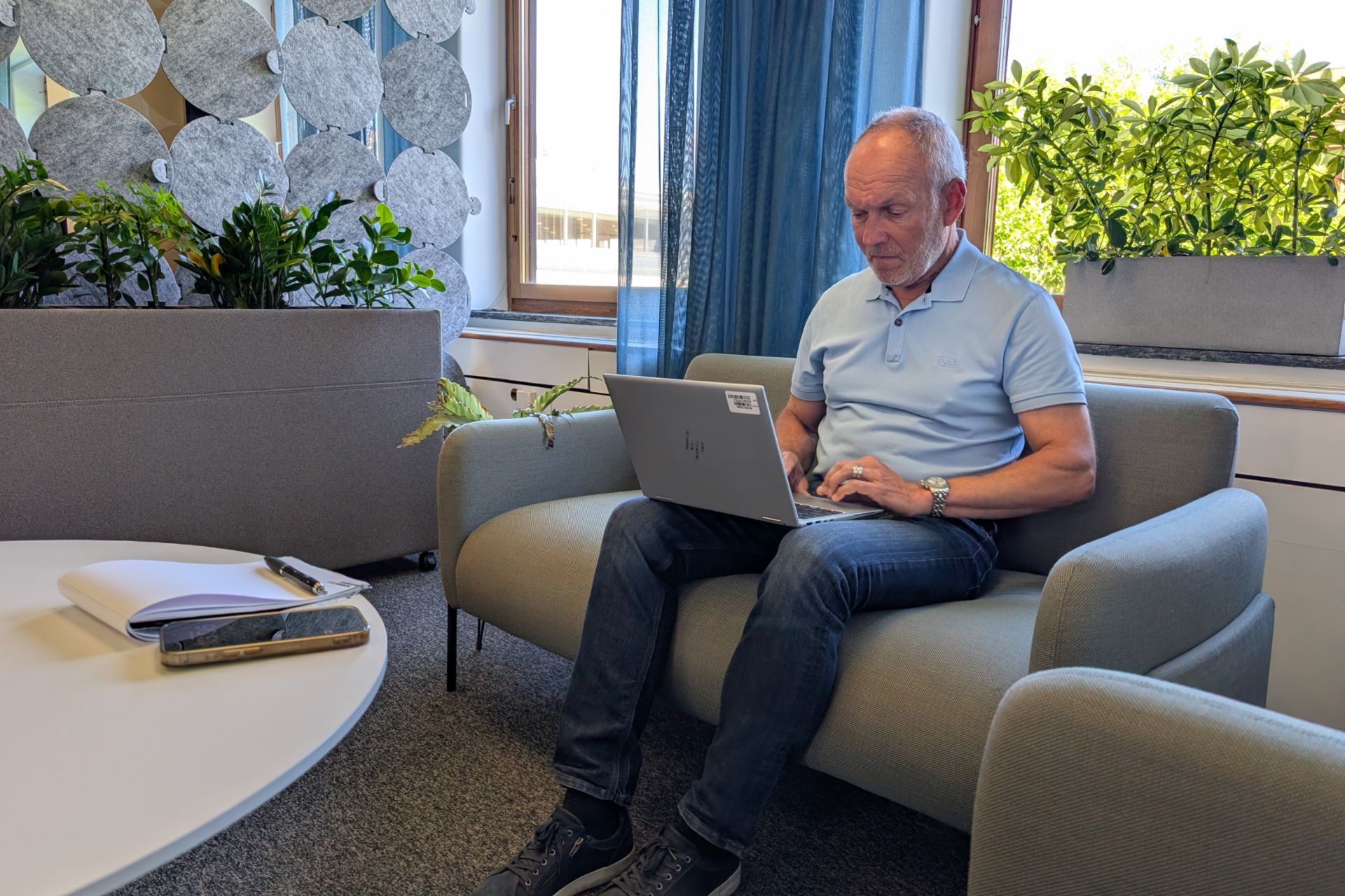  Ein Mann mit grauem Haar, bekleidet mit einem hellblauen Poloshirt und Jeans, sitzt auf einem grauen Sofa und tippt auf einem Laptop. Die Umgebung ist ein helles, modernes Büro mit Pflanzen und einer dekorativen Akustikwand aus grauen Filzkreisen. Im Vordergrund steht ein weißer Tisch, auf dem ein Notizbuch, ein Stift und ein Smartphone liegen.