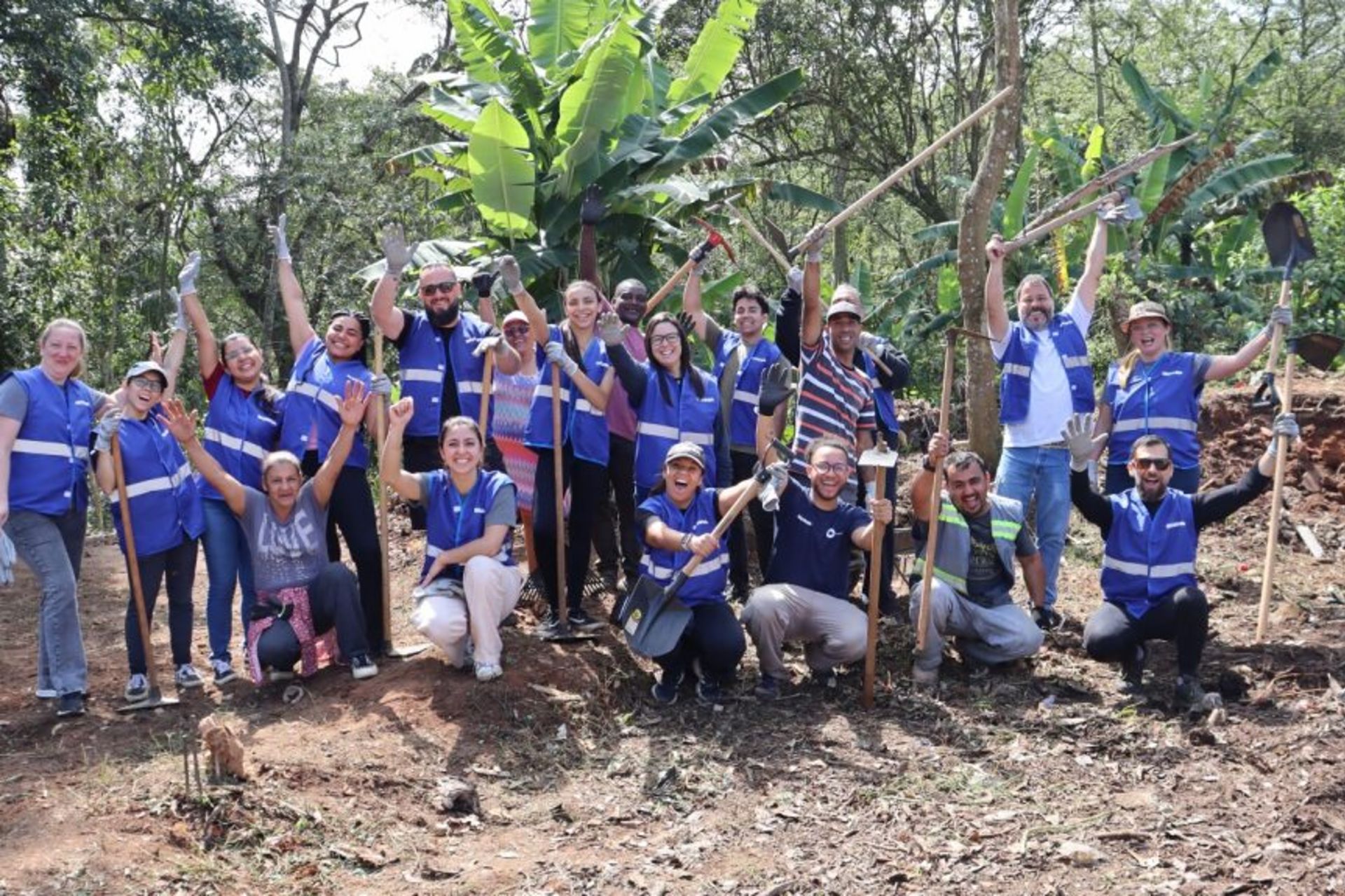 Several people are standing and kneeling in an open area within a wooded setting, holding shovels, hoes, and other tools raised above their heads. Many are wearing blue vests with reflective strips. Behind the group are banana plants, trees, and dense vegetation. The ground appears recently worked, suggesting gardening, reforestation, or landscape maintenance activities. The group has gathered for a photo at the work site.