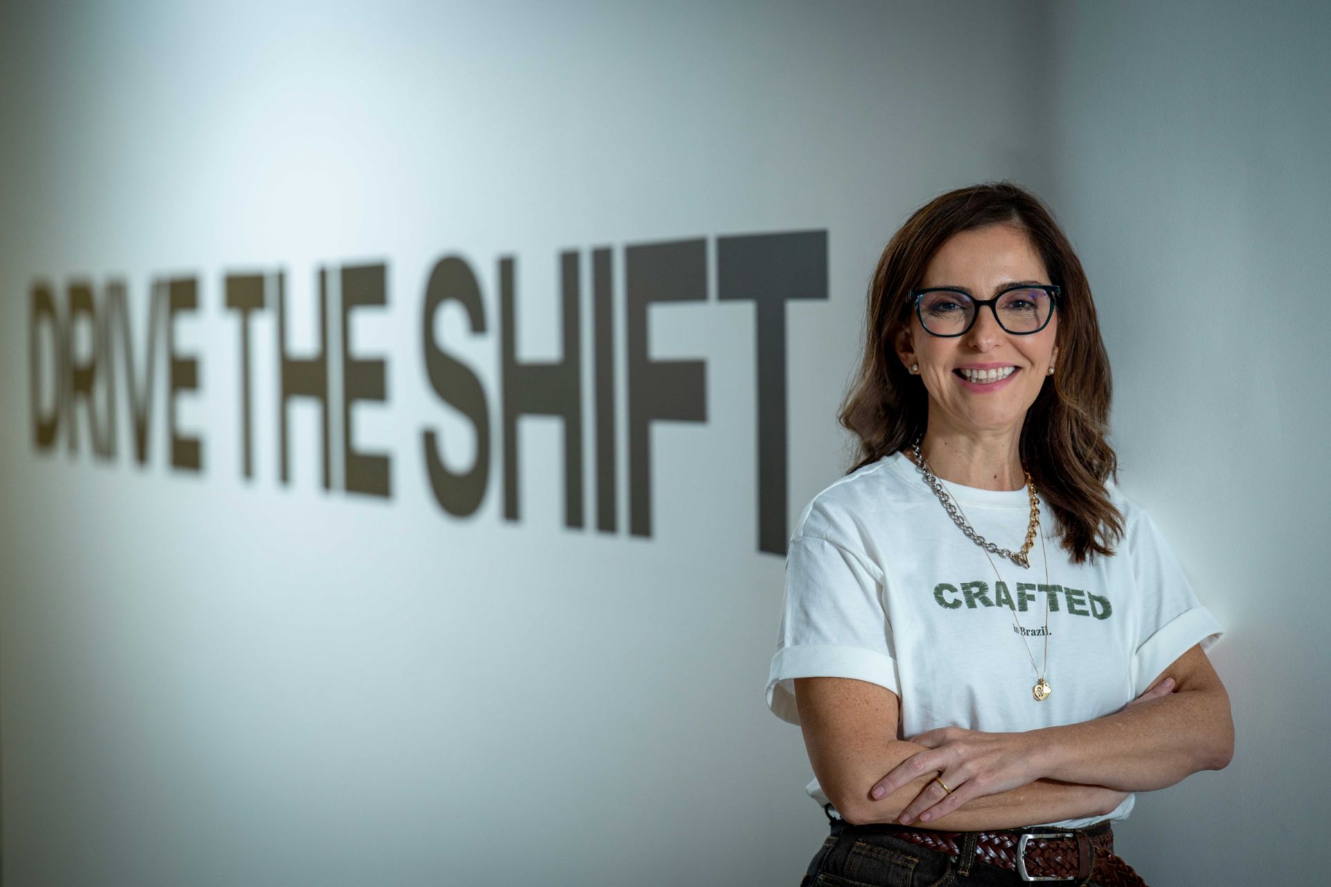 Patricia Acioli stands with arms crossed in front of a light-colored wall featuring the large text “DRIVE THE SHIFT.” Patricia Acioli is wearing glasses, a light-colored T-shirt displaying the words “CRAFTED in Brazil.”, and a necklace. The image shows Patricia Acioli from the waist up in an indoor setting with a clean background and prominent typographic design element.