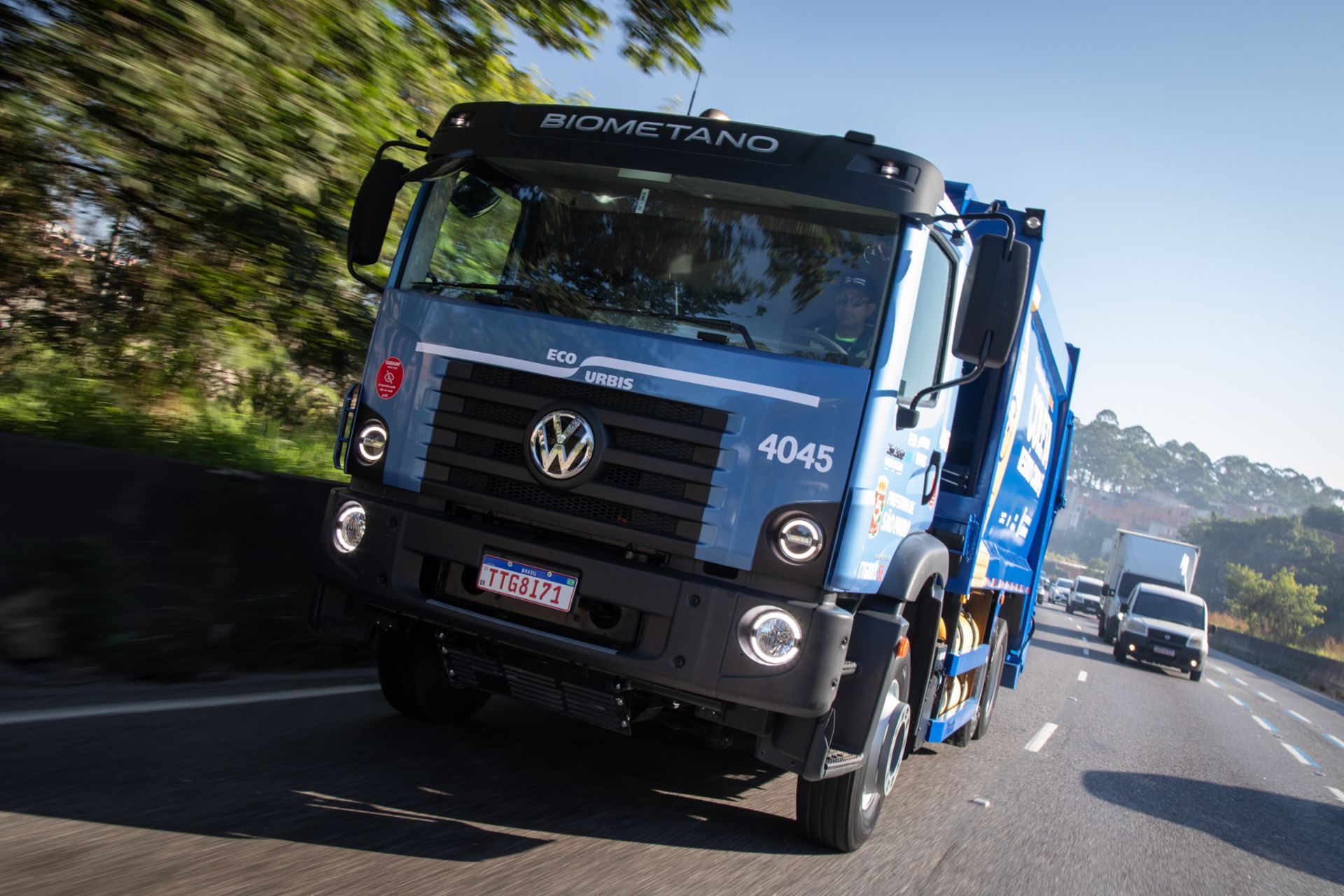 Blauer Volkswagen-Müllwagen fährt auf einer mehrspurigen Straße; im Hintergrund folgen weitere Fahrzeuge.