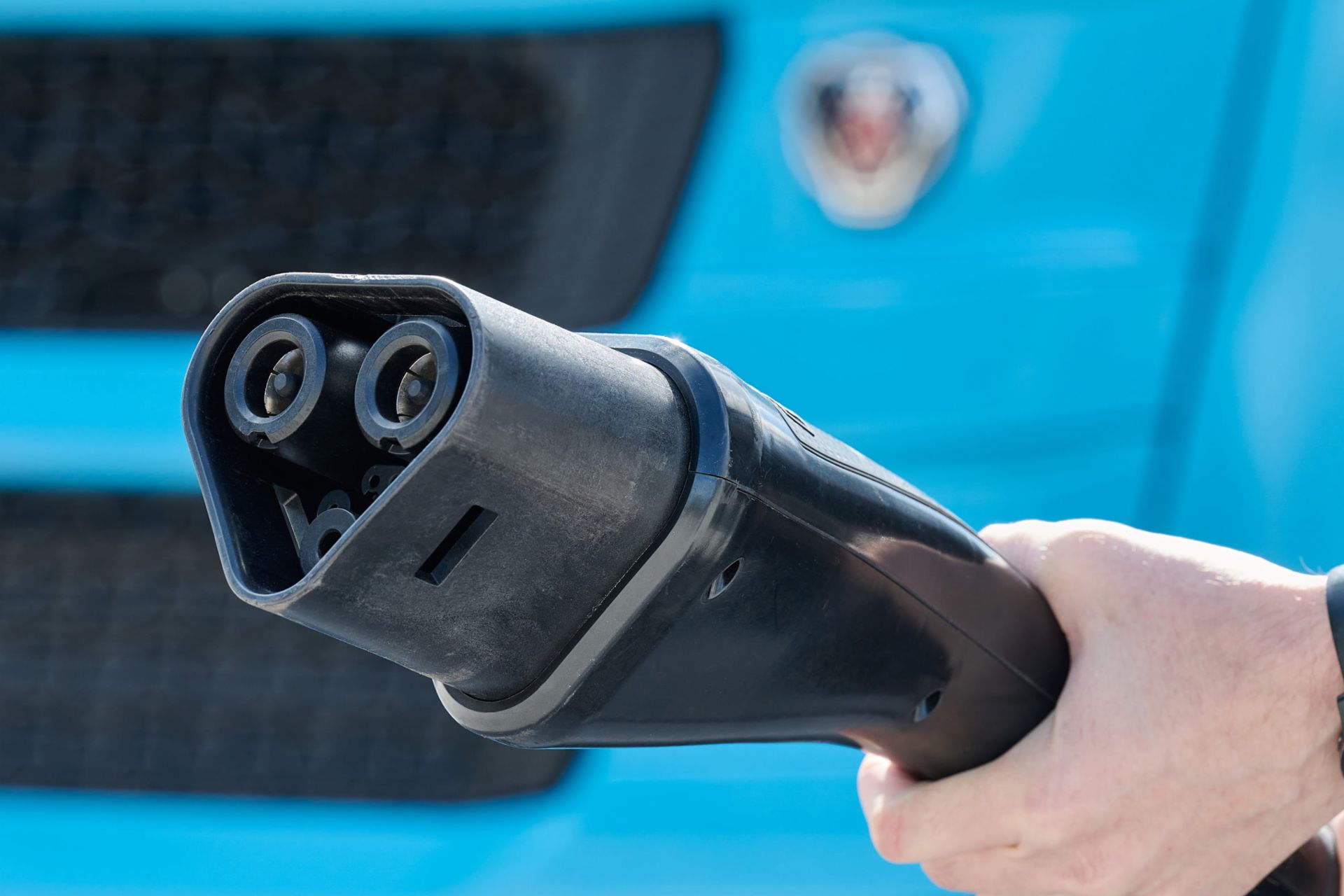 Nahaufnahme eines MCS-Ladesteckers (Megawatt Charging System), der von einer Person vor der Front eines blauen Elektro-Lkw gehalten wird.
                 