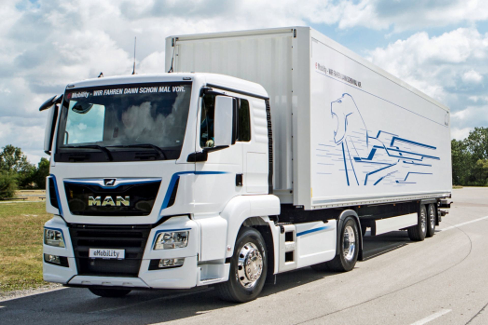 A fully electric MAN tractor unit with a dry-cargo trailer drives on a highway with blue skies in the background 
                 